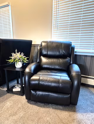 A black leather recliner chair positioned on a carpeted floor next to a small black side table with a white vase containing purple and green flowers. The background features two windows with white blinds and a beige wall with a gray wainscoting panel.