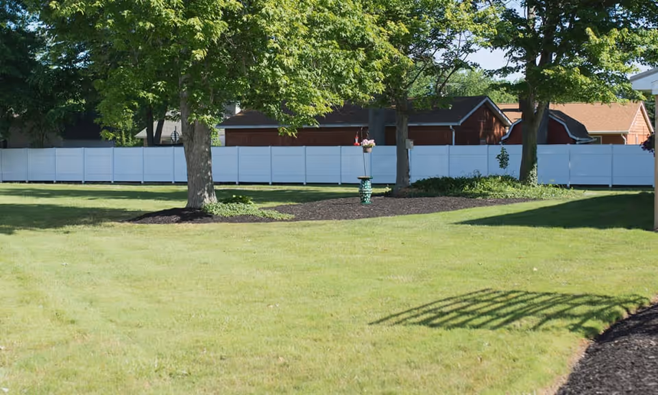 A grassy outdoor area with several trees and a white fence in the background. There is a small garden bed with mulch around the base of the trees and a decorative green bird feeder or planter in the center. Residential buildings with brown and beige siding are visible behind the fence.