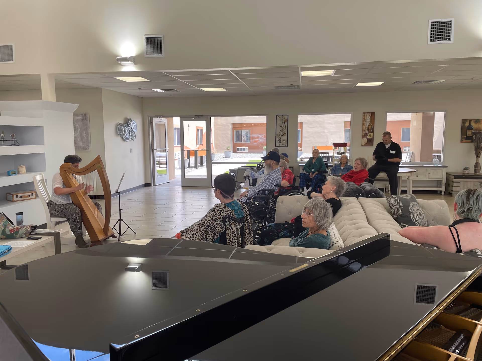 A group of elderly people seated on couches and chairs in a spacious, well-lit common area listening to a woman playing a harp. The room has large windows and a piano in the foreground.