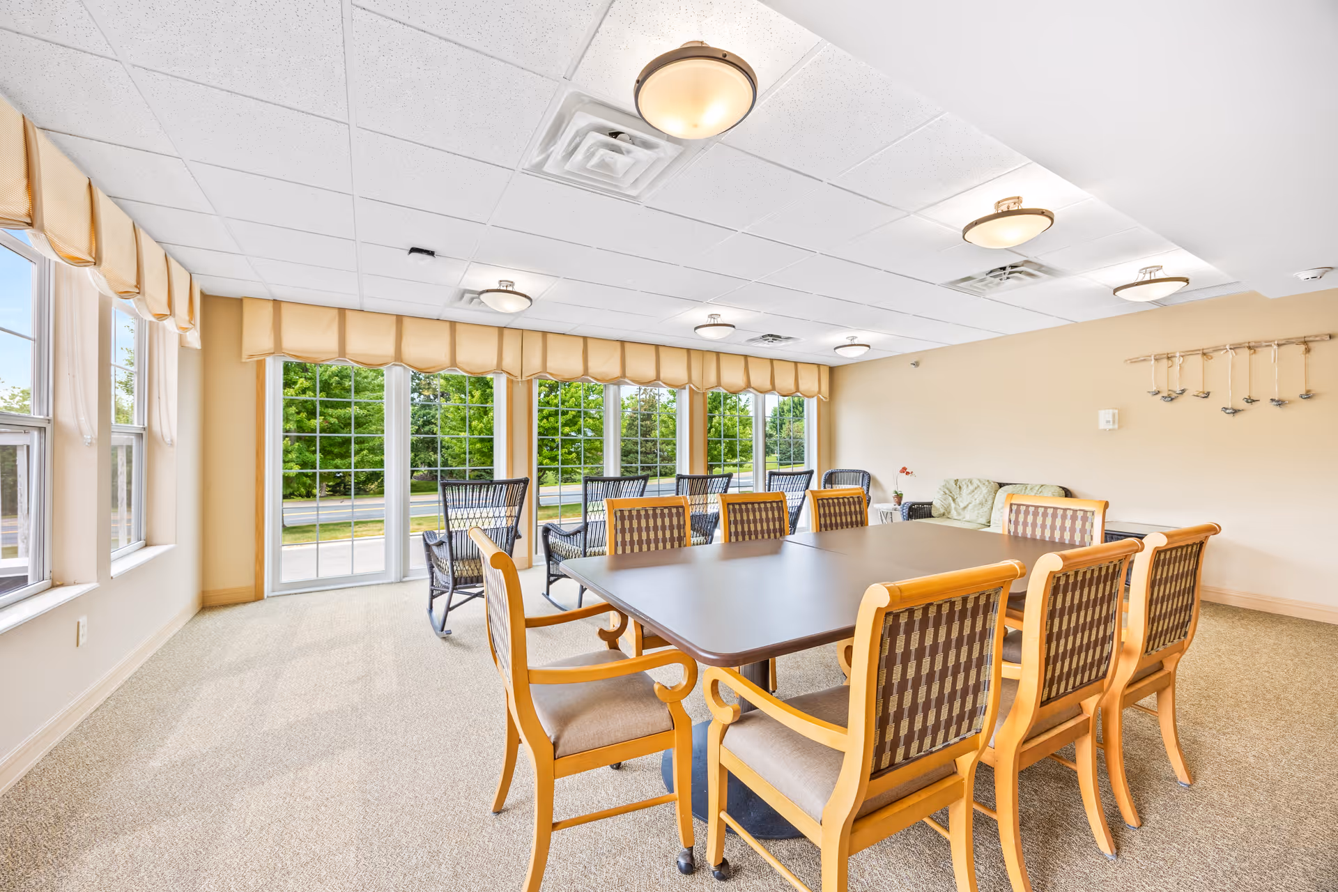 Bright common dining room with a long table surrounded by wooden chairs and large windows overlooking greenery.