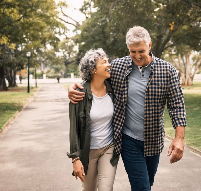 An older couple walking arm in arm down a tree-lined paved path in a park, smiling and enjoying each other's company on a sunny day.