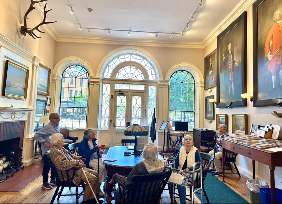 A bright lounge with tall arched windows, large portraits on the walls, a fireplace, a central table and several seated senior residents.
