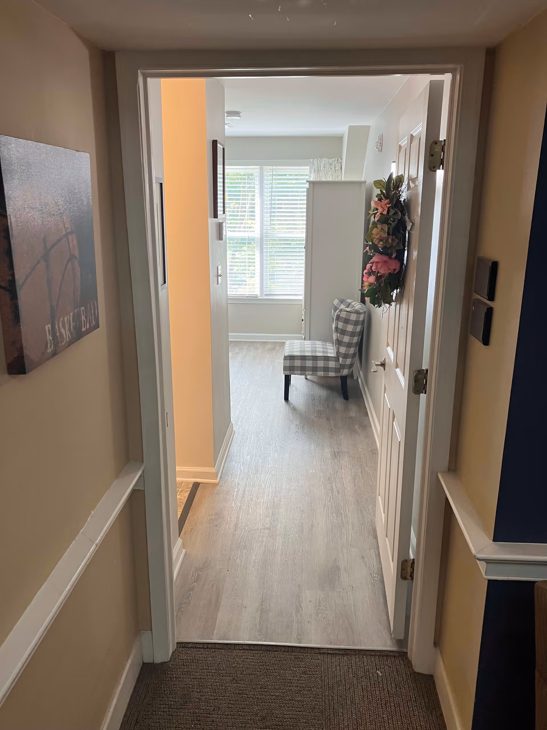 View through a doorway into a bright room with a large window covered by white blinds. A checkered upholstered chair is positioned near the window, and a floral wreath hangs on the door to the right. The floor is light wood, and the walls are painted in neutral tones. A basketball-themed picture is hung on the left wall in the hallway leading to the room.