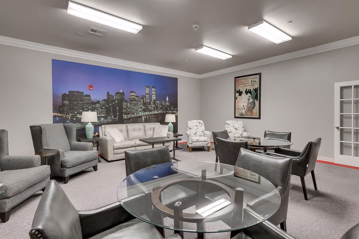 A senior living facility common area with multiple seating options including a beige sofa, gray armchairs, and patterned armchairs. There are two round glass tables with black leather chairs around them. The walls are light gray with a large cityscape mural and a framed vintage poster. The room is well-lit with fluorescent ceiling lights and has a carpeted floor.