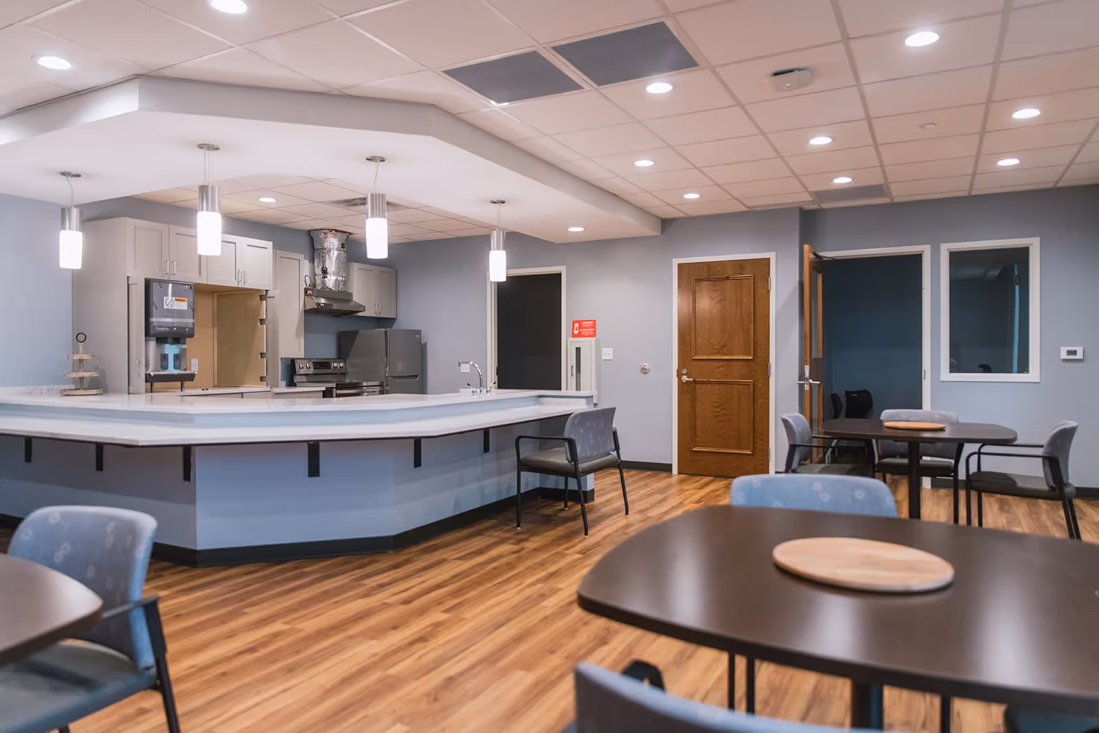 Interior view of a senior living facility dining area with a kitchen counter, several tables and chairs, wooden flooring, and ceiling lights. The kitchen area has cabinets, a stove, a refrigerator, and a paper towel dispenser. There are doors and windows on the walls.