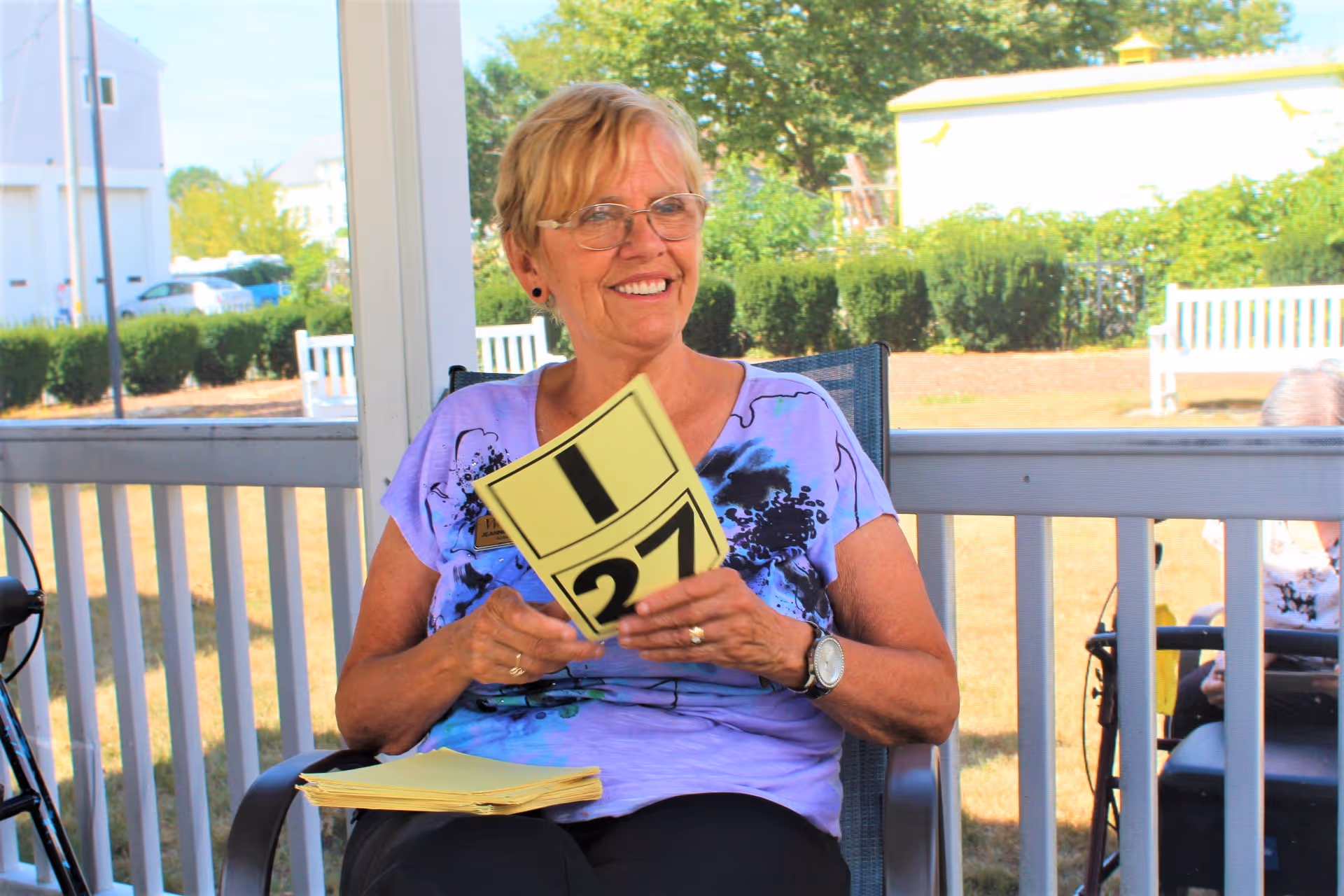 A smiling elderly woman wearing glasses and a purple shirt sits on a chair outdoors on a porch or patio, holding yellow cards with numbers on them. There is greenery and a white fence in the background.