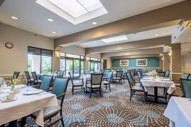 A spacious dining room with multiple tables covered in white tablecloths, each set with cups, plates, and small flower vases. The room features large windows allowing natural light to enter, patterned carpet flooring, and a ceiling with recessed lighting and skylights. The walls are decorated with framed artwork and a clock.