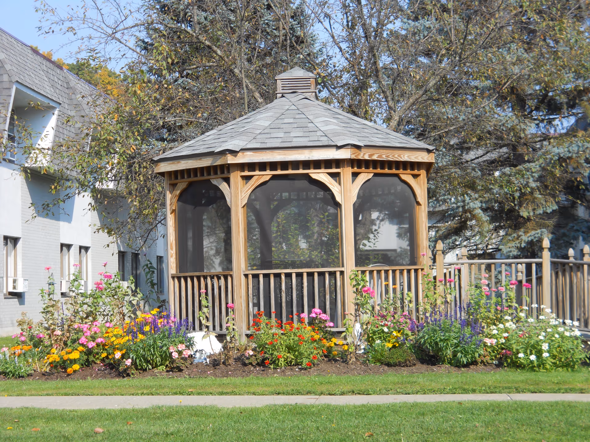 A wooden gazebo with a shingled roof and screened windows surrounded by a colorful flower garden. Behind the gazebo, there are trees and a building with white walls and windows.