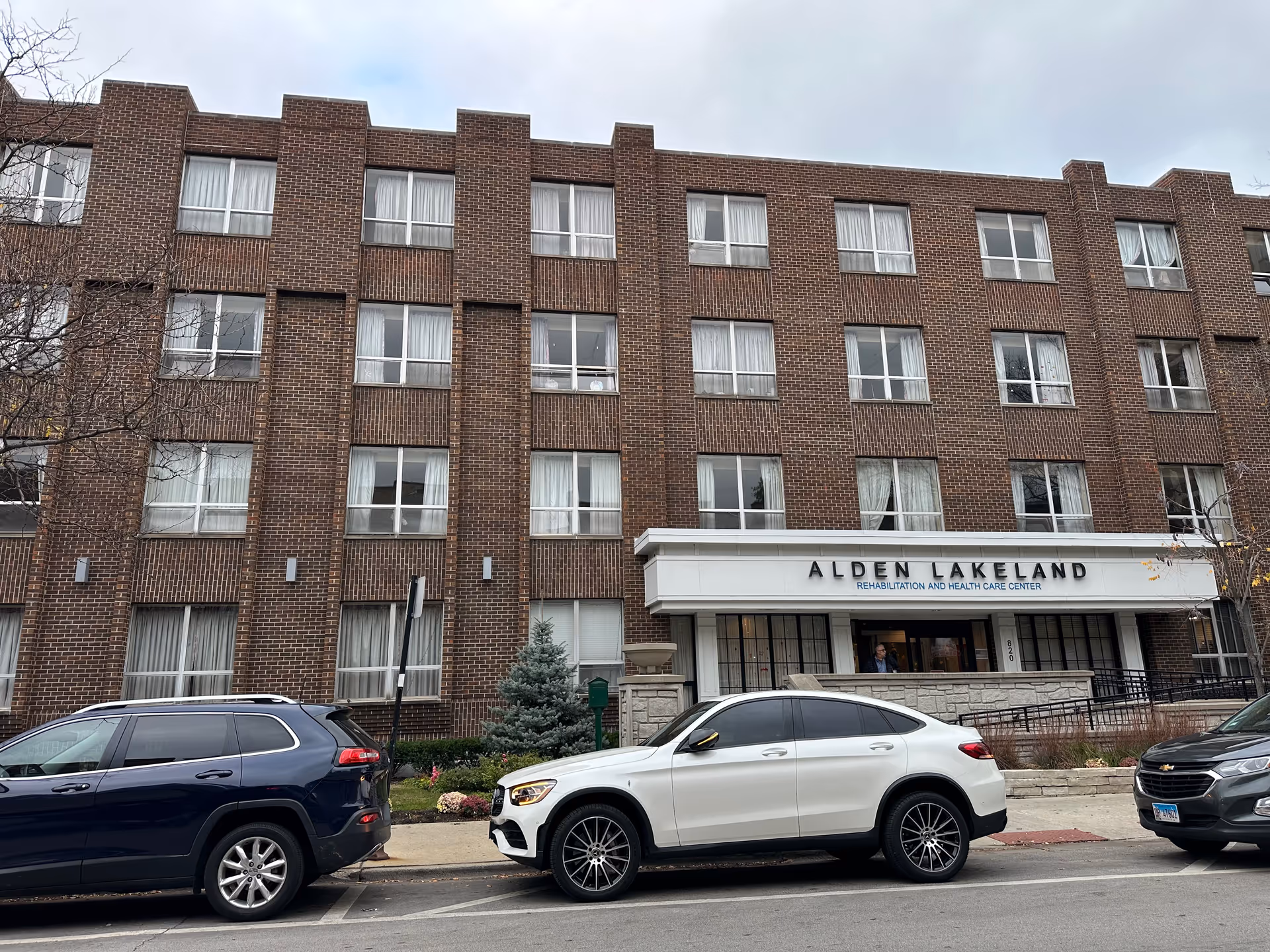 Front exterior view of a multi-story brick building with several windows and a white entrance canopy displaying the sign 'Alden Lakeland Rehabilitation and Health Care Center'. Three cars are parked along the street in front of the building.