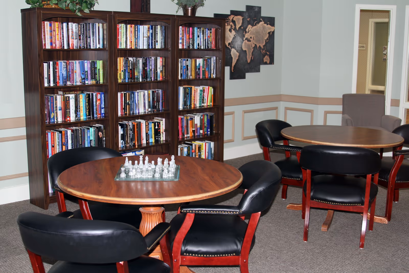 A cozy common room with round wooden tables surrounded by black chairs, a bookshelf filled with books, a chess set on one table, and a world map on the wall.