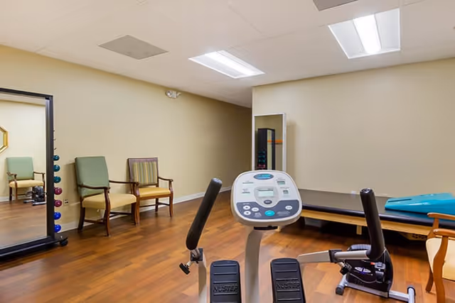 A small exercise room with wooden flooring, featuring a stationary exercise bike in the foreground, two chairs against the wall, a large mirror on the left, and a padded exercise bench with a blue mat on it in the background.