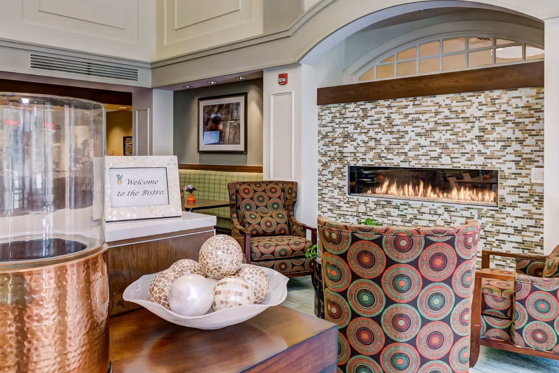 A cozy seating area in a bistro with patterned armchairs arranged around a modern rectangular fireplace set in a tiled wall. A wooden table in the foreground holds a decorative bowl with mosaic balls and a sign that reads 'Welcome to the Bistro.' The background shows a booth seating area with a framed picture on the wall.