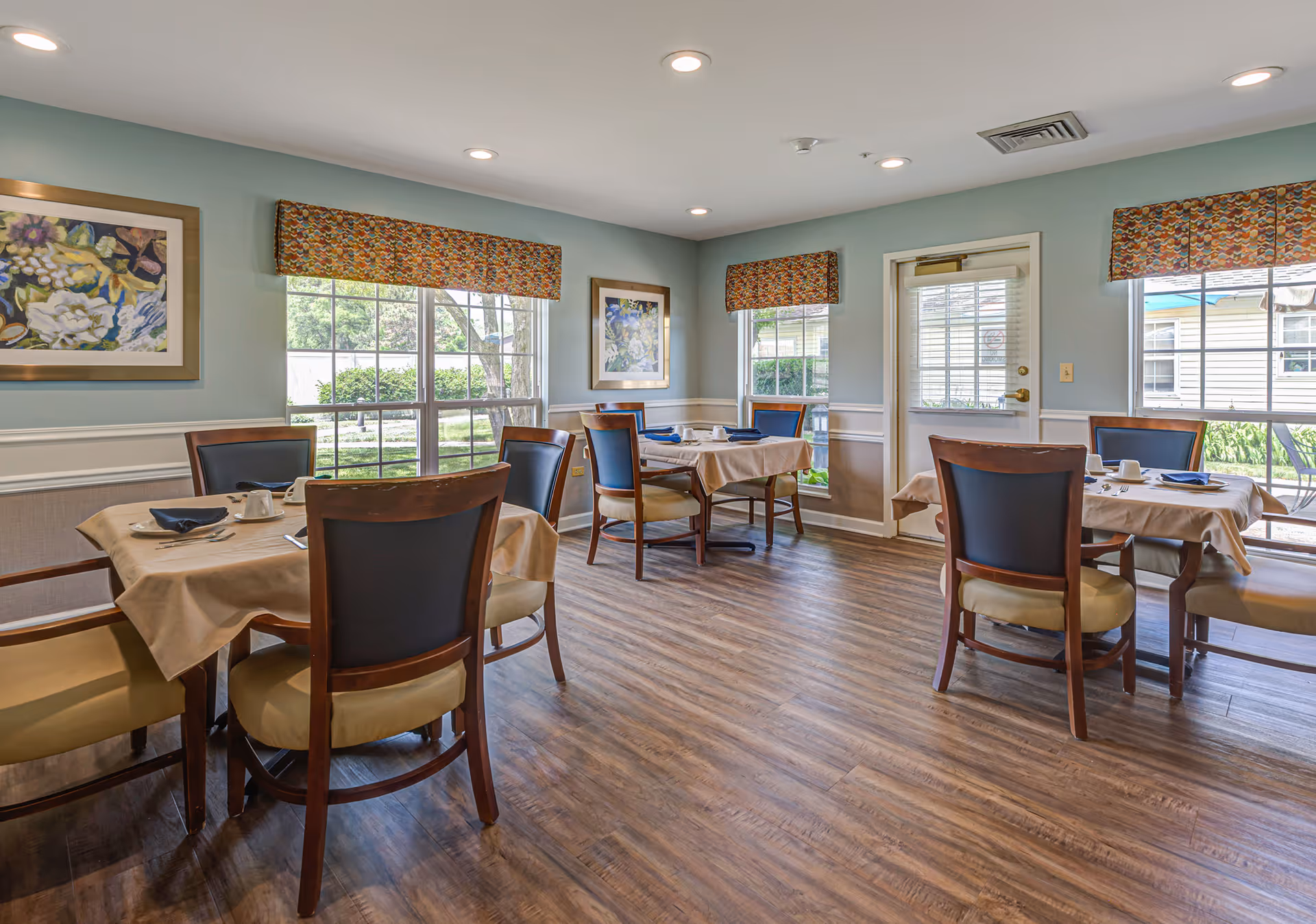 A dining room in a senior living facility with several tables covered with beige tablecloths, each set with plates, cups, and napkins. The room has wooden floors, light blue walls with white wainscoting, large windows with colorful valances, and framed floral artwork on the walls. There is a door leading outside and natural light fills the room.