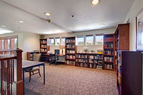 A well-lit library or reading room with multiple wooden bookshelves filled with books, a desk with a computer and chair, a table with chairs, and windows letting in natural light.