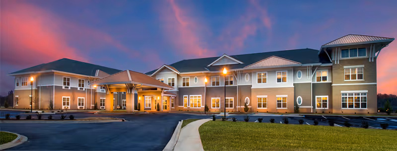 Exterior view of a two-story senior living facility building at dusk with a lit entrance canopy, multiple windows, and a paved driveway surrounded by landscaped grass areas under a colorful sky.