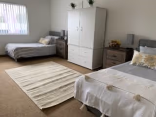 A bright bedroom with two beds, each with pillows and blankets. Between the beds is a nightstand with a lamp and decorative items. A white wardrobe with drawers stands against the wall, and a light-colored rug is placed on the carpeted floor. A window with vertical blinds allows natural light into the room.