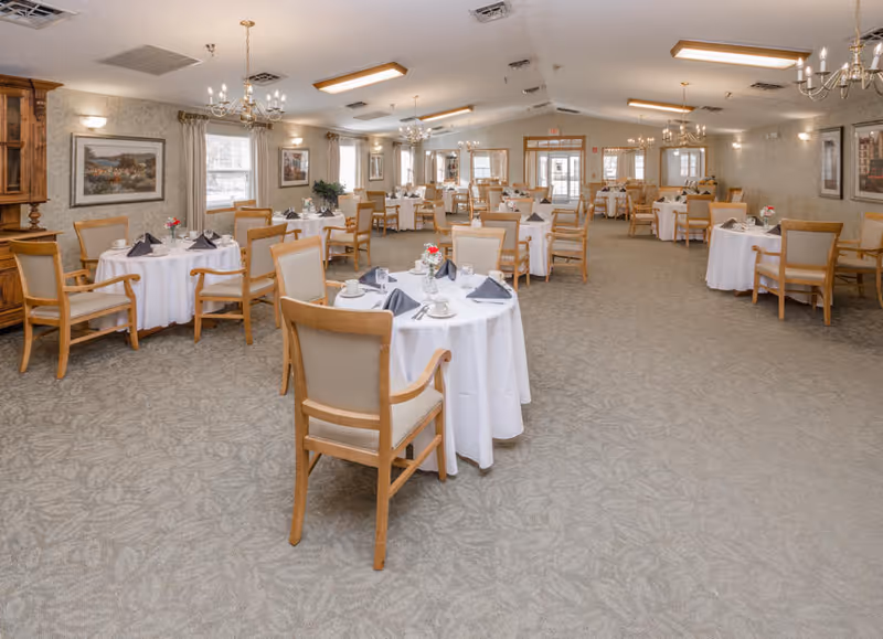 A spacious dining room with round tables covered in white tablecloths, place settings and wooden chairs arranged under chandeliers.