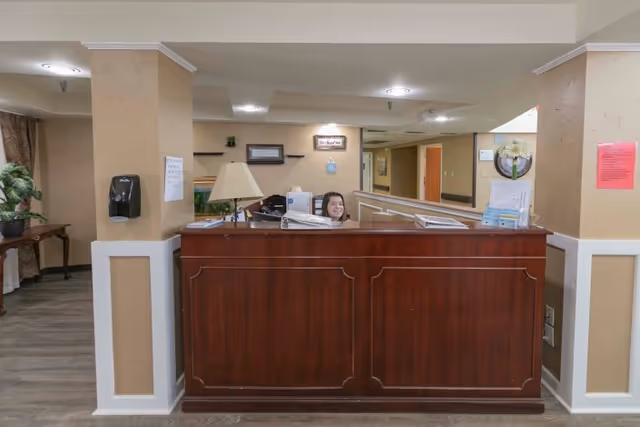 Reception desk inside a senior living facility with a staff member smiling behind the wooden counter.