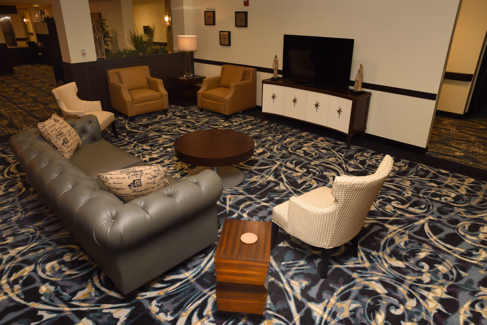 A cozy living room area with a gray tufted sofa, two tan armchairs, and two beige patterned chairs arranged around a round wooden coffee table. There is a wooden side table with a coaster, a TV on a white and brown cabinet, and a lamp on a side table. The floor is covered with a patterned carpet in shades of blue, beige, and black.