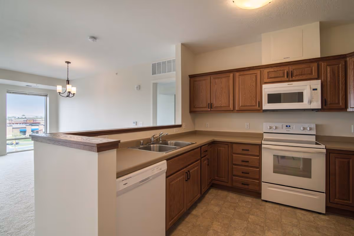 Interior view of a kitchen with wooden cabinets, a white stove, microwave, dishwasher, and a double sink. The kitchen opens into a dining area with a hanging light fixture and a large window showing an outdoor view.