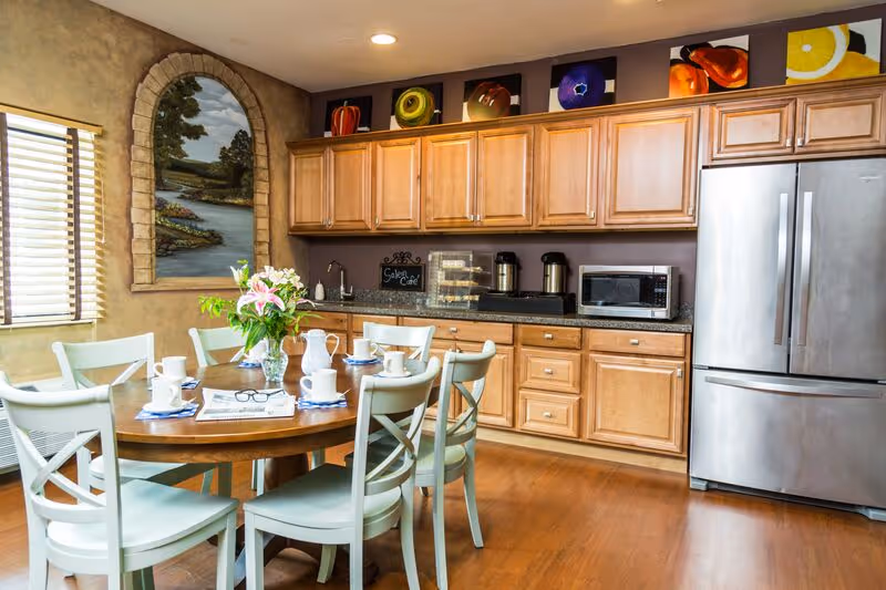 A bright kitchen and dining area with wooden cabinets, a stainless steel refrigerator, a round wooden table surrounded by six light green chairs, and a vase of flowers on the table. The wall features a decorative mural of a landscape and colorful fruit paintings above the cabinets.