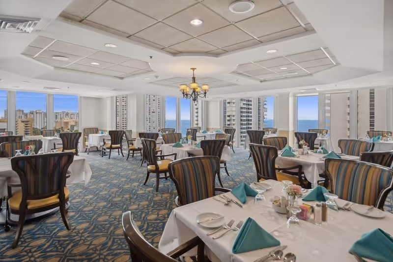 A bright and elegant dining room with large windows offering a view of tall buildings and the ocean. The room features multiple round tables covered with white tablecloths, set with plates, silverware, glasses, and teal folded napkins. The chairs have dark wooden frames with striped upholstery. A chandelier hangs from the ceiling, and the carpet has a blue and beige pattern.