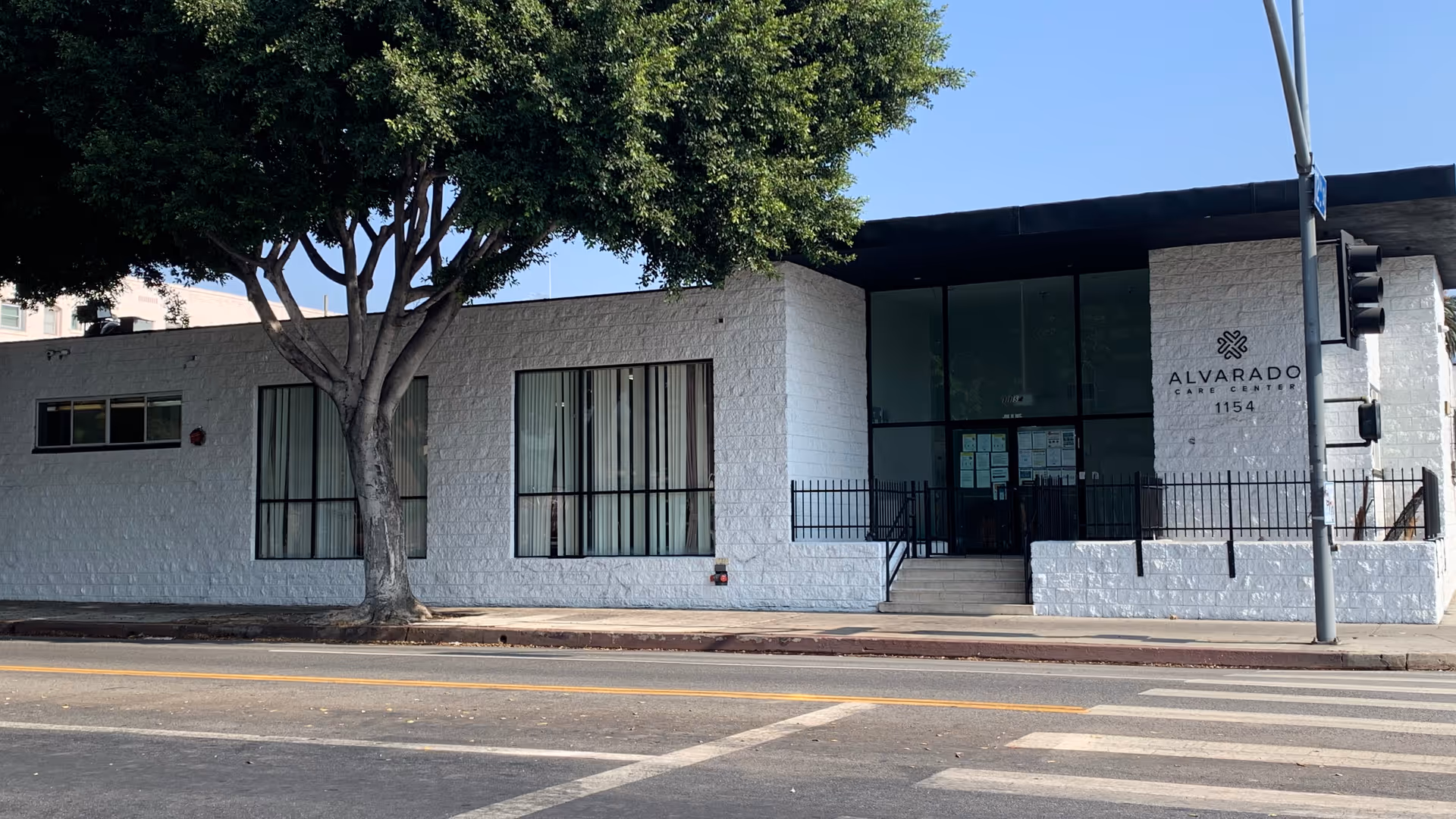 Exterior view of Alvarado Care Center, a single-story white building with large windows and a glass entrance door. There is a tree in front of the building and a pedestrian crosswalk on the street in front.
