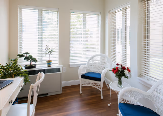 Sun-filled seating area with white wicker chairs and blue cushions around a small table with a vase of red flowers, potted plants, and large windows with blinds.