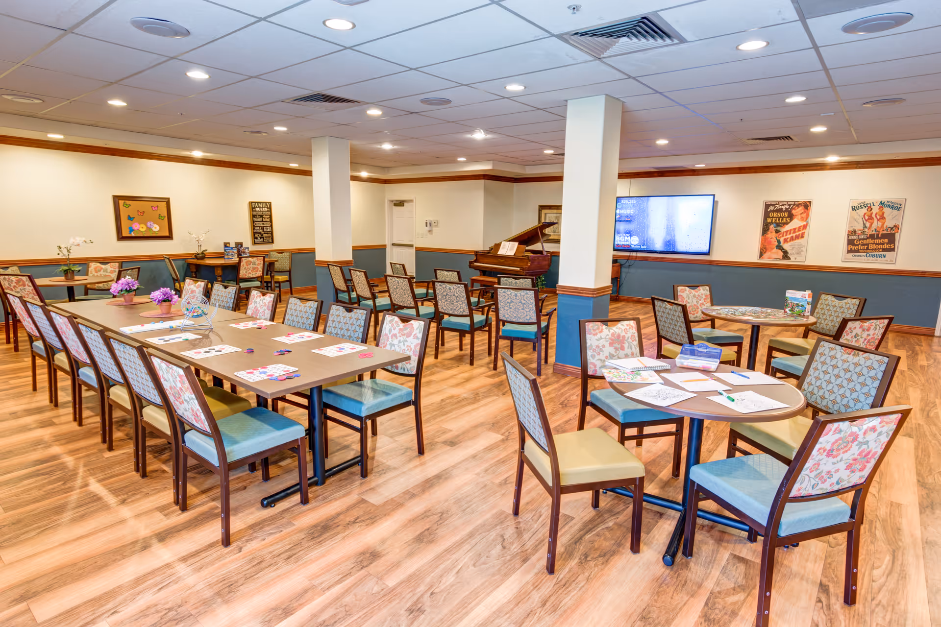Bright communal dining and activity room with multiple tables and chairs, a piano, a wall-mounted TV, and activity materials on the tables.