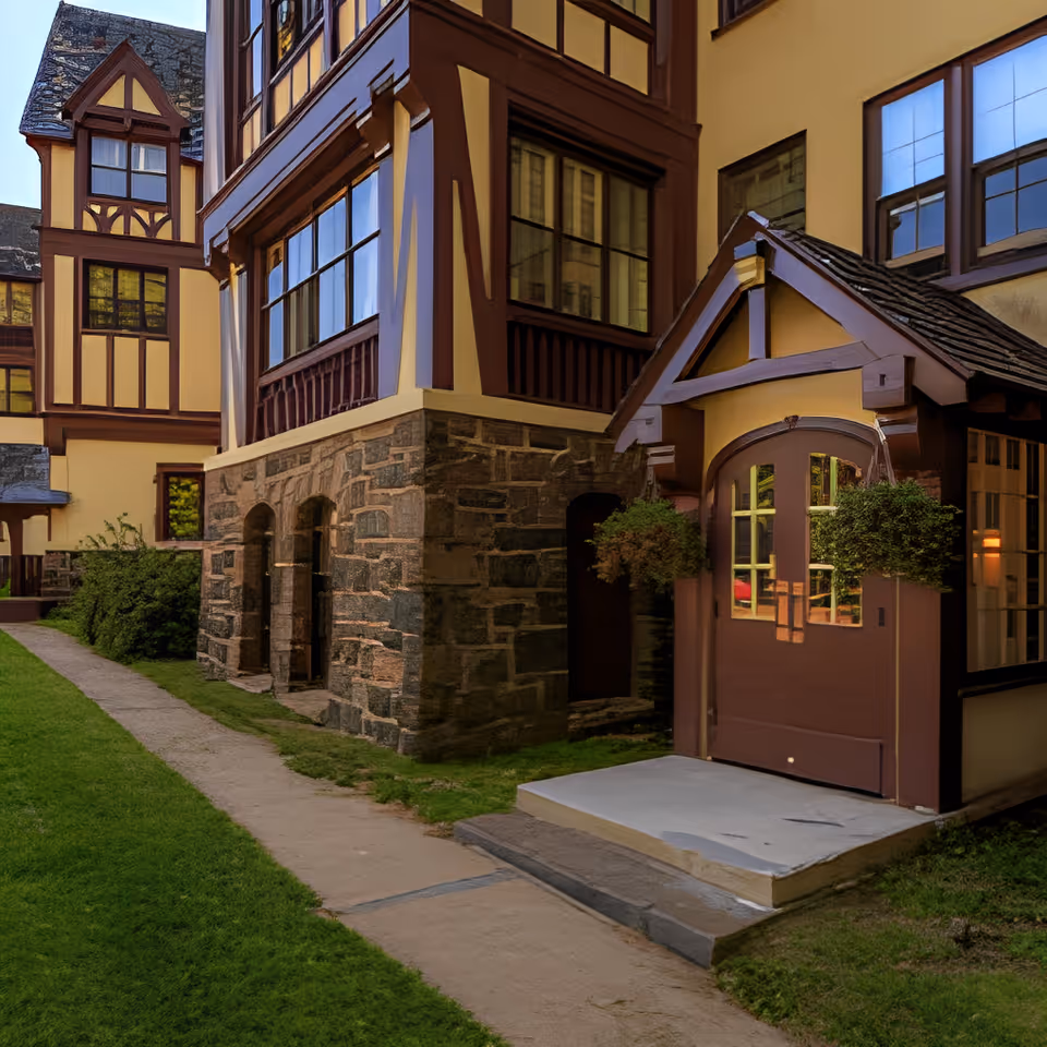 Tudor-style building exterior with a grass courtyard path, stone foundation, and a small covered wooden entrance with hanging planters.