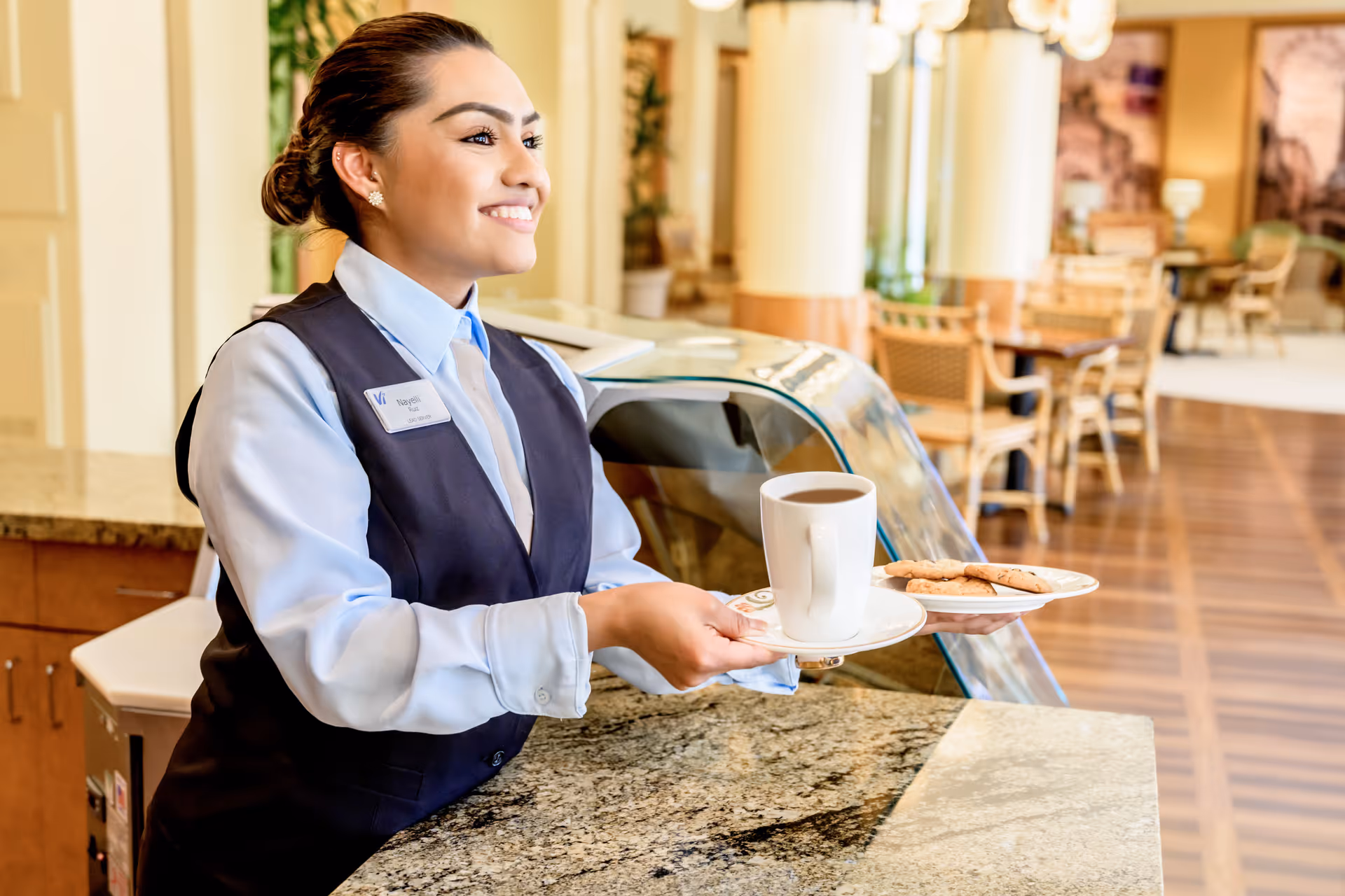 A smiling female staff member wearing a uniform and name tag stands behind a granite counter in a well-lit dining area, holding a tray with a cup of coffee and a plate of cookies. The background shows tables and chairs arranged in the dining space with warm lighting and decor.