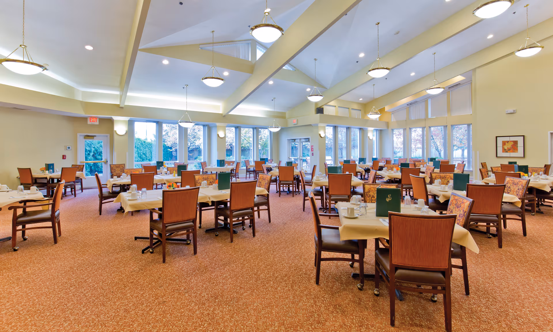 A spacious dining room with multiple tables covered in beige tablecloths, each set with cups, glasses, and menus. The room has large windows letting in natural light, high ceilings with hanging light fixtures, and a patterned carpeted floor. Chairs with wooden frames and cushioned seats surround the tables.