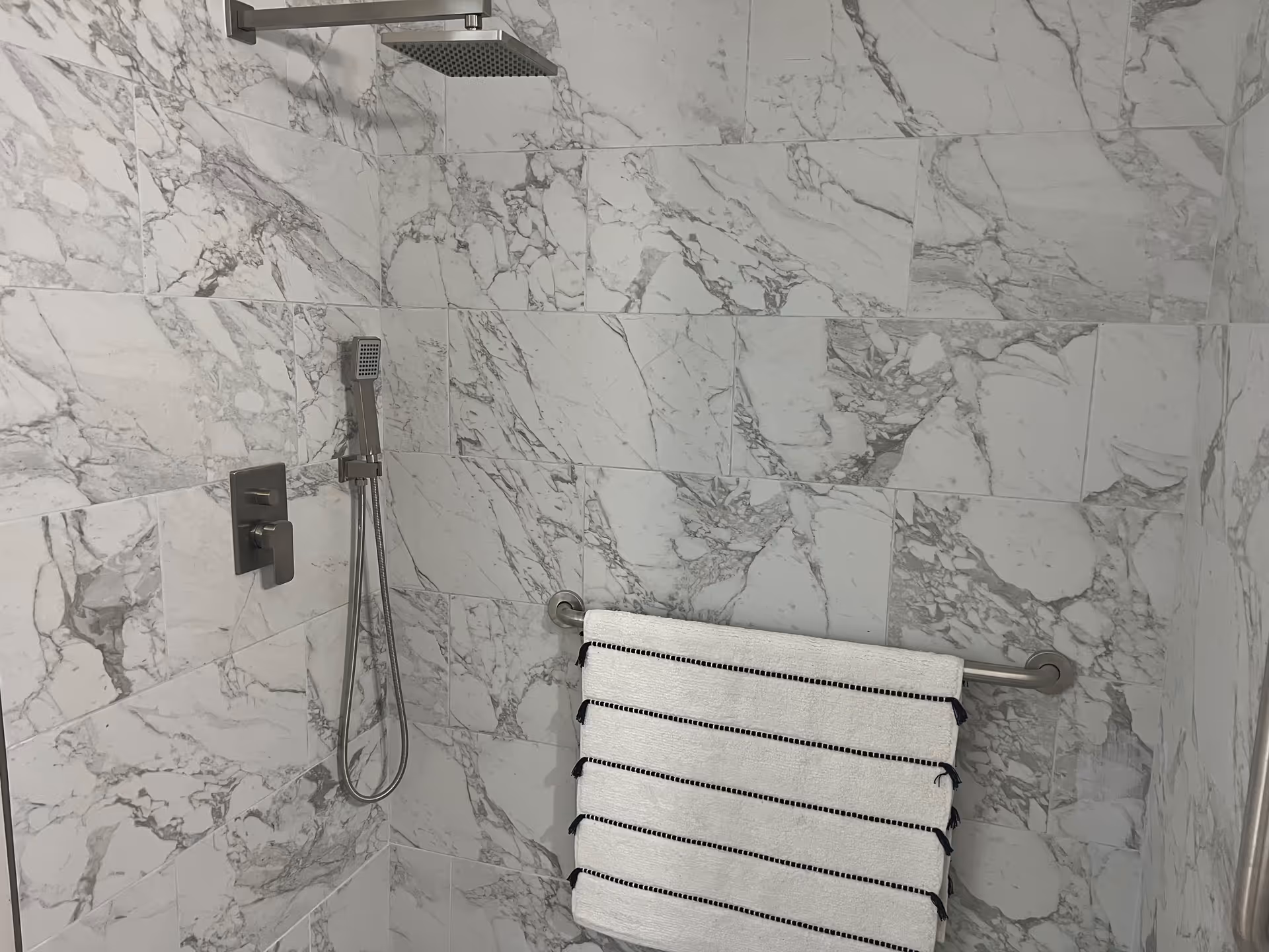 A modern shower area with white and gray marble tiles covering the walls. The shower features a square overhead rain showerhead, a handheld showerhead, and a control panel. A white towel with black stripes is hanging on a metal towel bar attached to the wall.