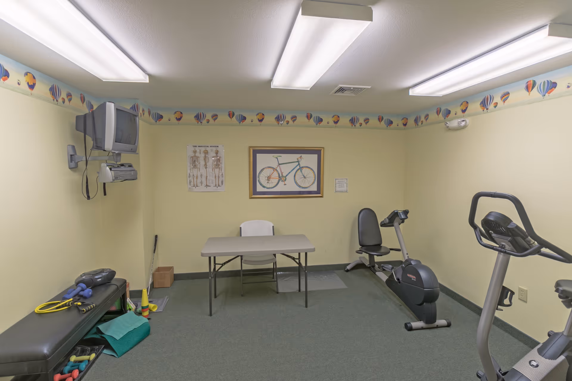 A small exercise room with light yellow walls and a hot air balloon border near the ceiling. The room contains a recumbent exercise bike, an upright exercise bike, a folding table with a white chair, a wall-mounted TV, and various exercise equipment including dumbbells and resistance bands on a bench. There are two framed pictures on the wall, one of a bicycle and one of a skeletal system diagram.