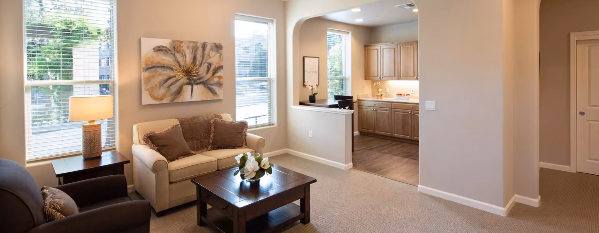 A cozy living room with a beige loveseat adorned with two brown pillows and a brown throw blanket, a dark brown armchair with a matching pillow, and a wooden coffee table with a flower arrangement. The room has two large windows with white blinds, a table lamp on a side table, and a large floral painting on the wall. An open archway leads to a kitchen area with wooden cabinets and a small dining nook with two chairs.