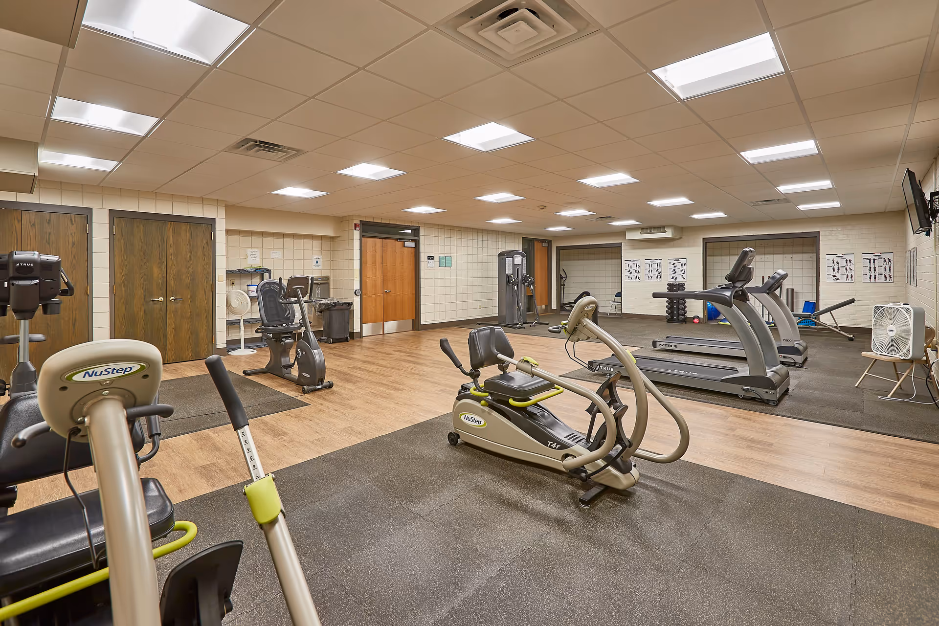 A well-lit fitness room with various exercise equipment including NuStep recumbent cross trainers, treadmills, and weight machines. The room has wood and rubber flooring, beige walls with exercise posters, and a wall-mounted TV. There are wooden double doors and a water fountain in the background.