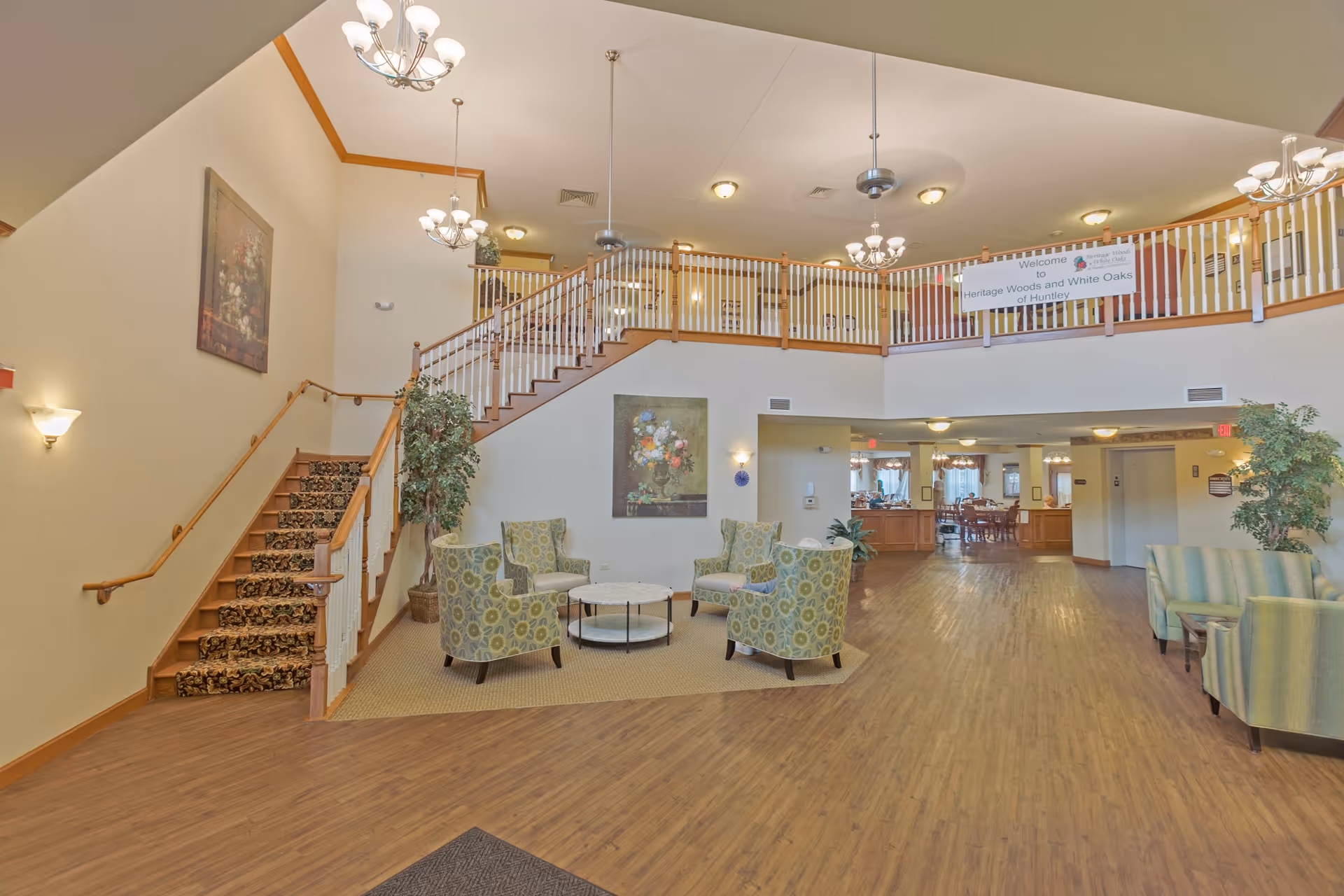 Spacious senior living facility common area with wooden flooring, a staircase with carpeted steps and wooden railing, several patterned armchairs arranged around a circular coffee table on a carpeted section, potted plants, and a balcony with a railing above. A banner on the balcony reads 'Welcome to Heritage Woods and White Oaks of Huntley'. The area is well-lit with multiple chandeliers and wall sconces.