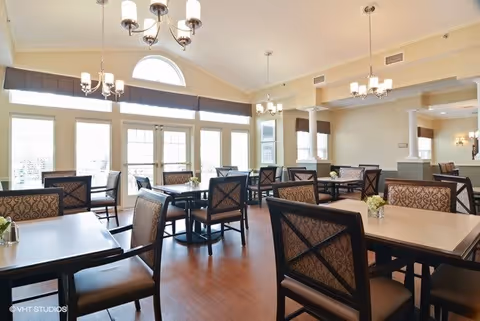 A bright and spacious dining room with multiple tables and chairs arranged neatly. Large windows with white frames and a half-circle window above allow natural light to fill the room. The walls are painted a light color, and there are several chandeliers hanging from the ceiling. Small flower arrangements are placed on the tables.
