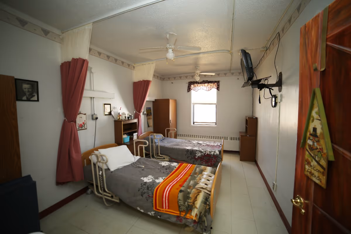 A simple room with two hospital-style beds, each with gray floral blankets and one with an additional orange and red striped blanket. The beds have metal side rails. The room has white walls with a decorative border near the ceiling, two ceiling fans, a window with a valance, and wooden furniture including a wardrobe and a small shelving unit. There are privacy curtains between the beds and a wall-mounted TV. A door with a 'Welcome Friends' sign is partially open.