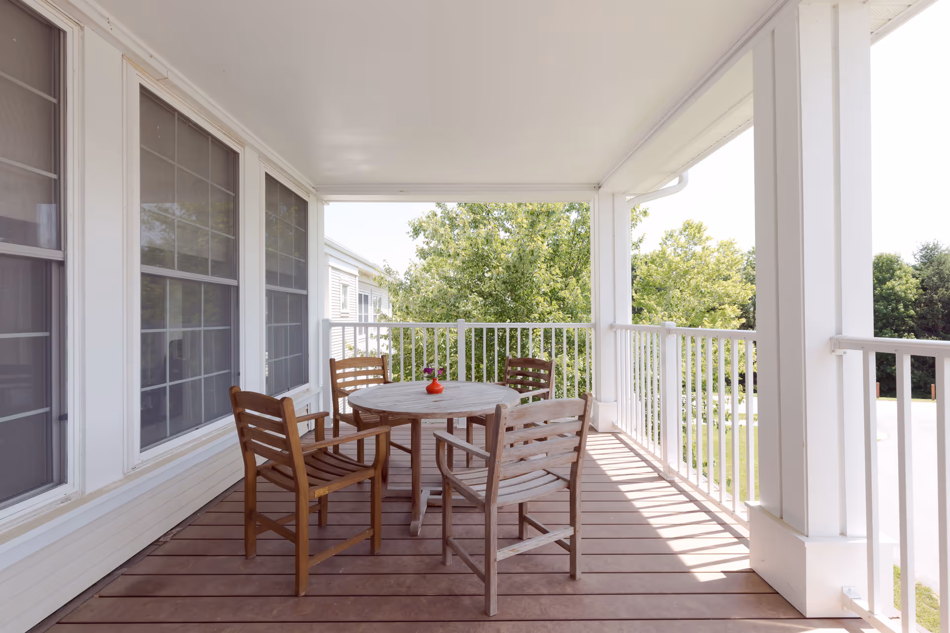 Covered outdoor balcony with a round wooden table and four wooden chairs. A small red vase with flowers is placed on the table. The balcony has white railings and overlooks green trees and a clear sky.