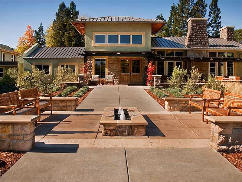 Outdoor seating area with wooden benches and stone planters surrounding a rectangular fire pit, leading to the entrance of a building with large windows and a metal roof, surrounded by trees and shrubs.