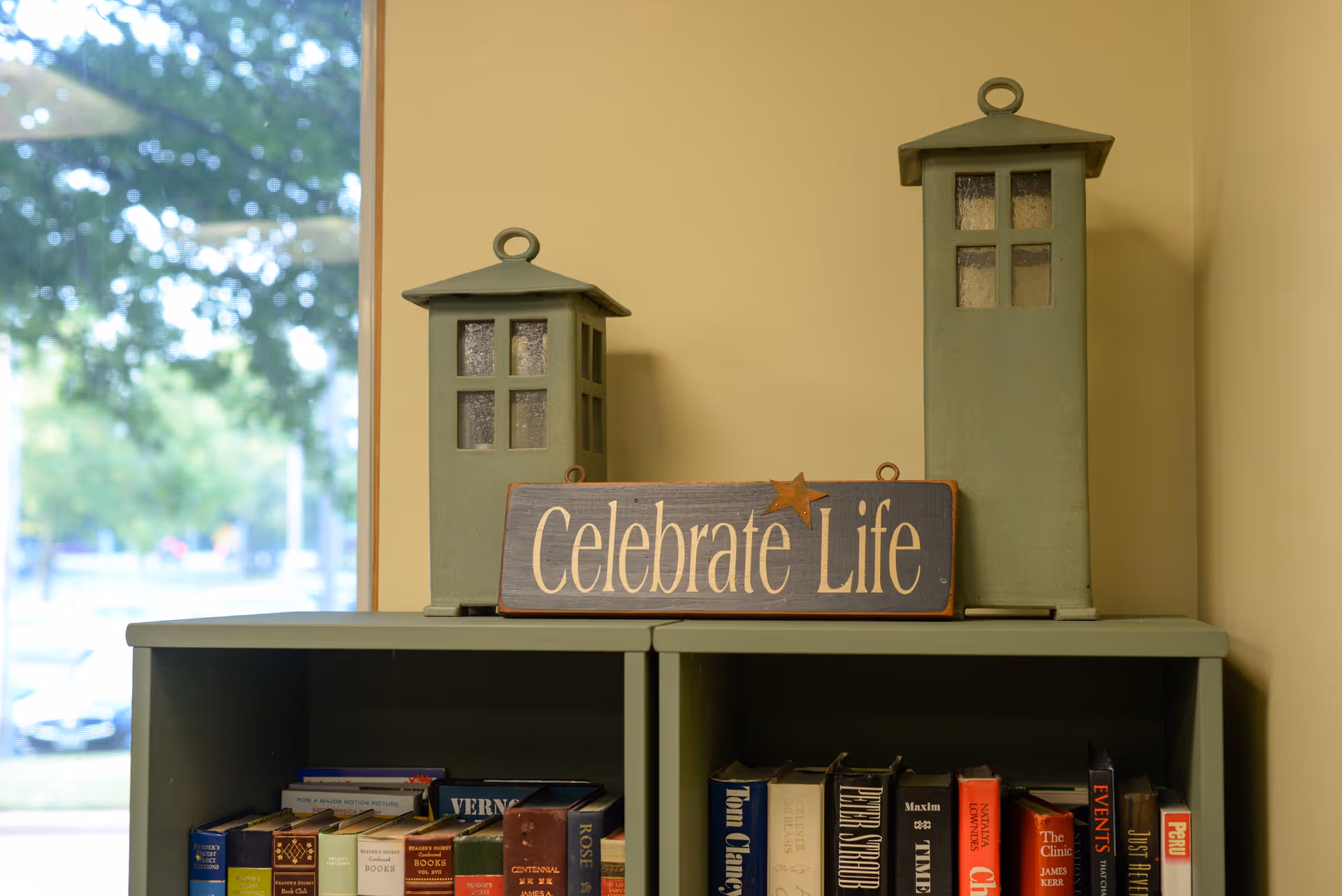 A green shelf topped with two decorative lanterns, a wooden 'Celebrate Life' sign, and books on the lower shelves beside a window.
