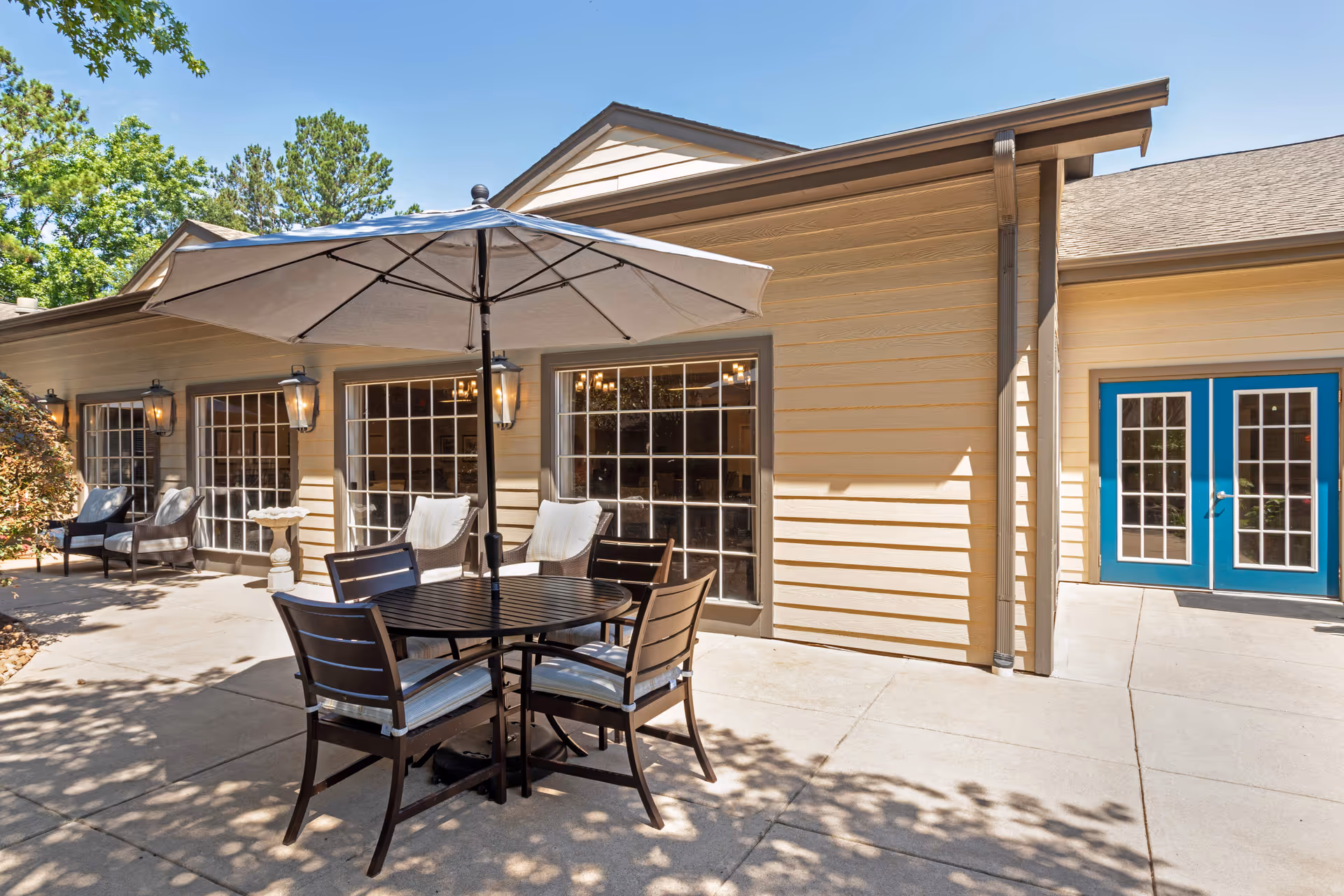 Outdoor patio area at Lewis Park Estates Senior Living with a round table, four chairs with cushions, and a large umbrella providing shade. The building exterior is beige with large windows and blue double doors. Trees and shrubs surround the patio area.