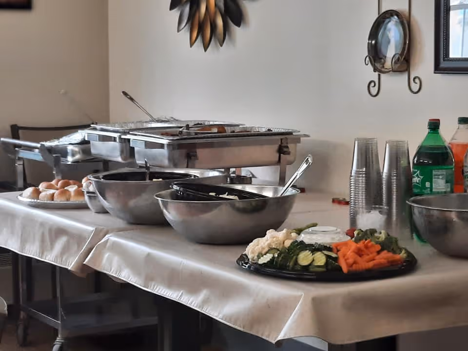 A buffet table set up with trays of food, bowls, a vegetable platter with dip, rolls, plastic cups, and bottles of soda in a dining area with wall decorations.
