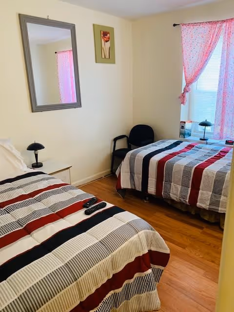 A small bedroom with two twin beds covered in striped bedspreads in shades of red, black, gray, and white. There is a black chair between the beds, a small nightstand with a black lamp, a wall mirror, and a framed flower picture on the wall. A window with pink floral curtains is on the right side, letting in natural light.