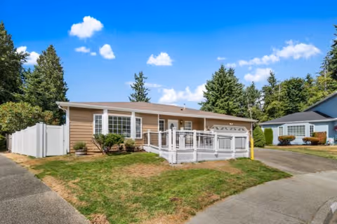 Single-story house with beige siding and white trim, featuring a white railing around a small front porch. The house is surrounded by a lawn with some bushes and trees in the background under a blue sky with scattered clouds.