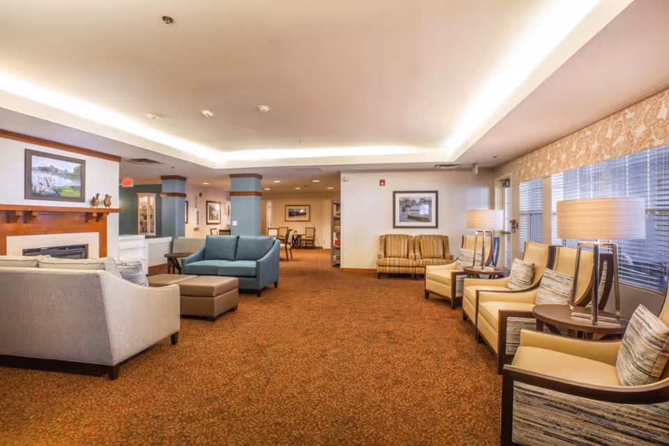 A spacious and well-lit living room area in a senior living facility featuring multiple seating arrangements including beige armchairs with cushions, a blue loveseat, a gray sofa, and a brown ottoman. The room has a patterned carpet, a fireplace with a wooden mantle, framed artwork on the walls, and large windows with blinds and valances. Several table lamps provide additional lighting.