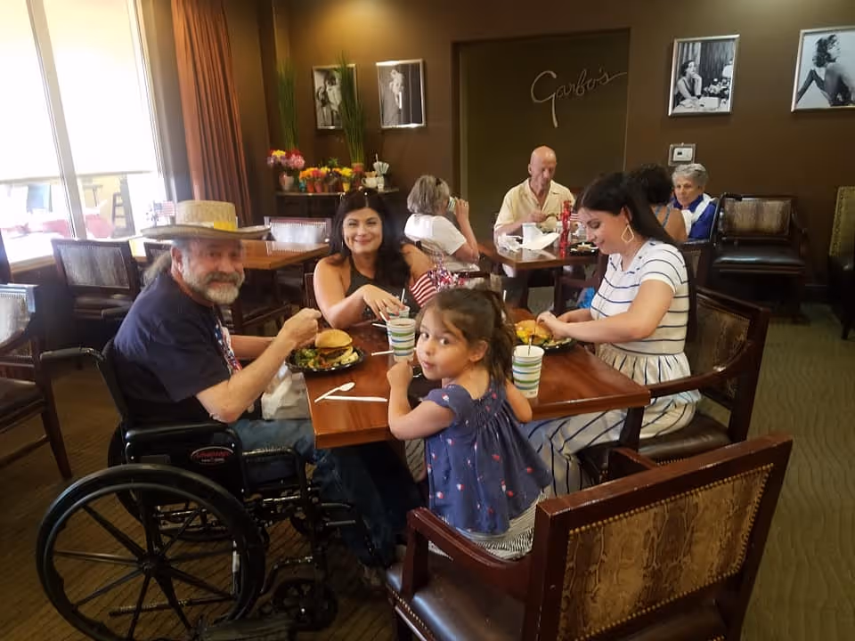Several people, including a man in a wheelchair, a young child, and adults, sitting around a dining table eating in a senior living dining room.
