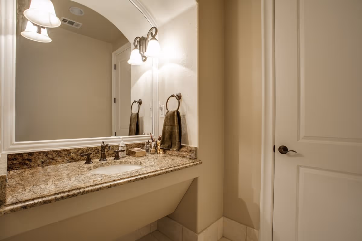 A bathroom vanity area with a granite countertop, an under-mount sink, bronze faucet fixtures, a large mirror with a white frame, and two wall-mounted light fixtures with frosted glass shades. A brown hand towel hangs on a bronze towel ring next to the sink, and a closed white door with a bronze handle is visible on the right side.