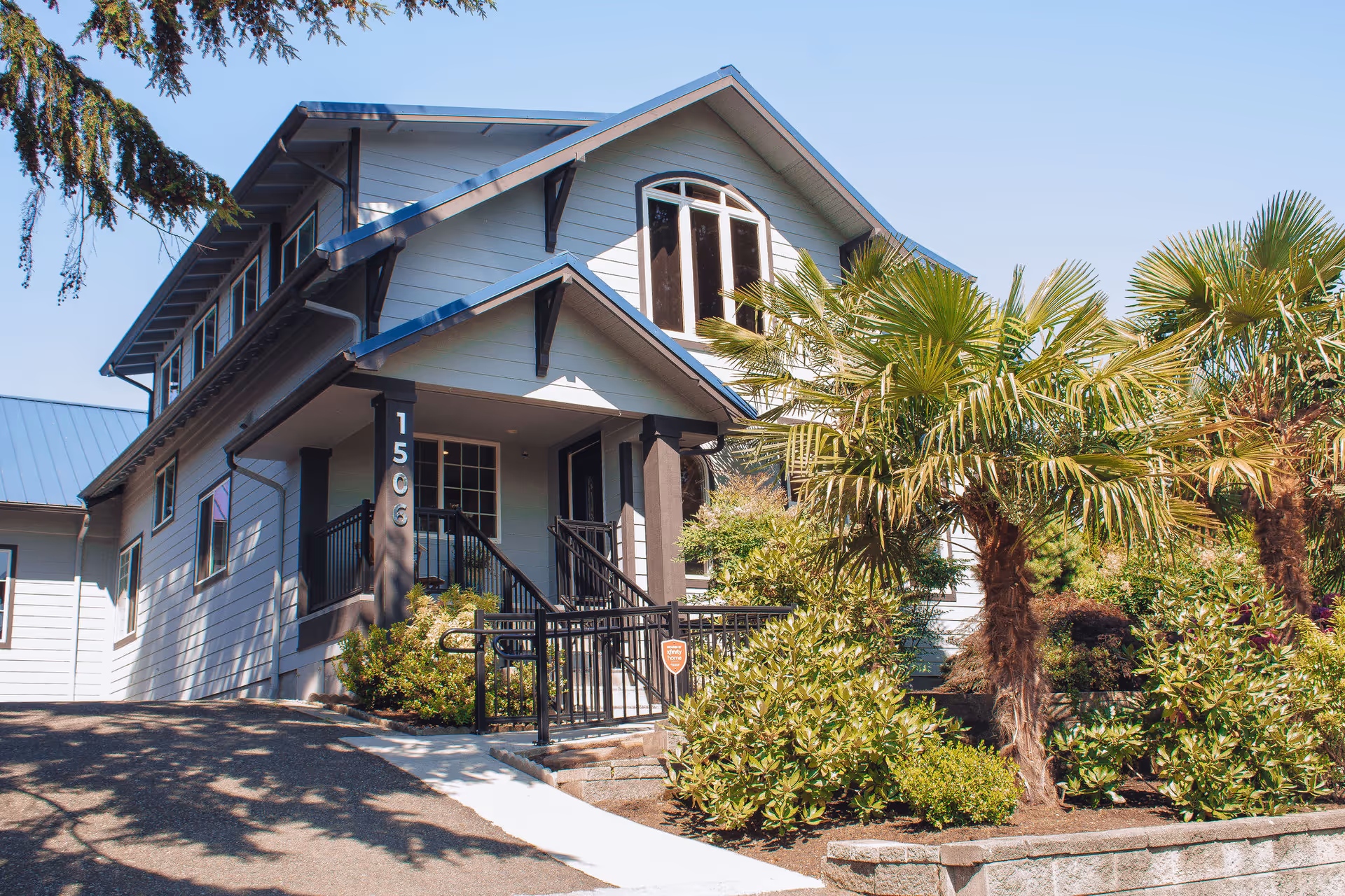 Two-story light-gray house with a covered front porch, wheelchair ramp, and landscaped yard with palm trees.